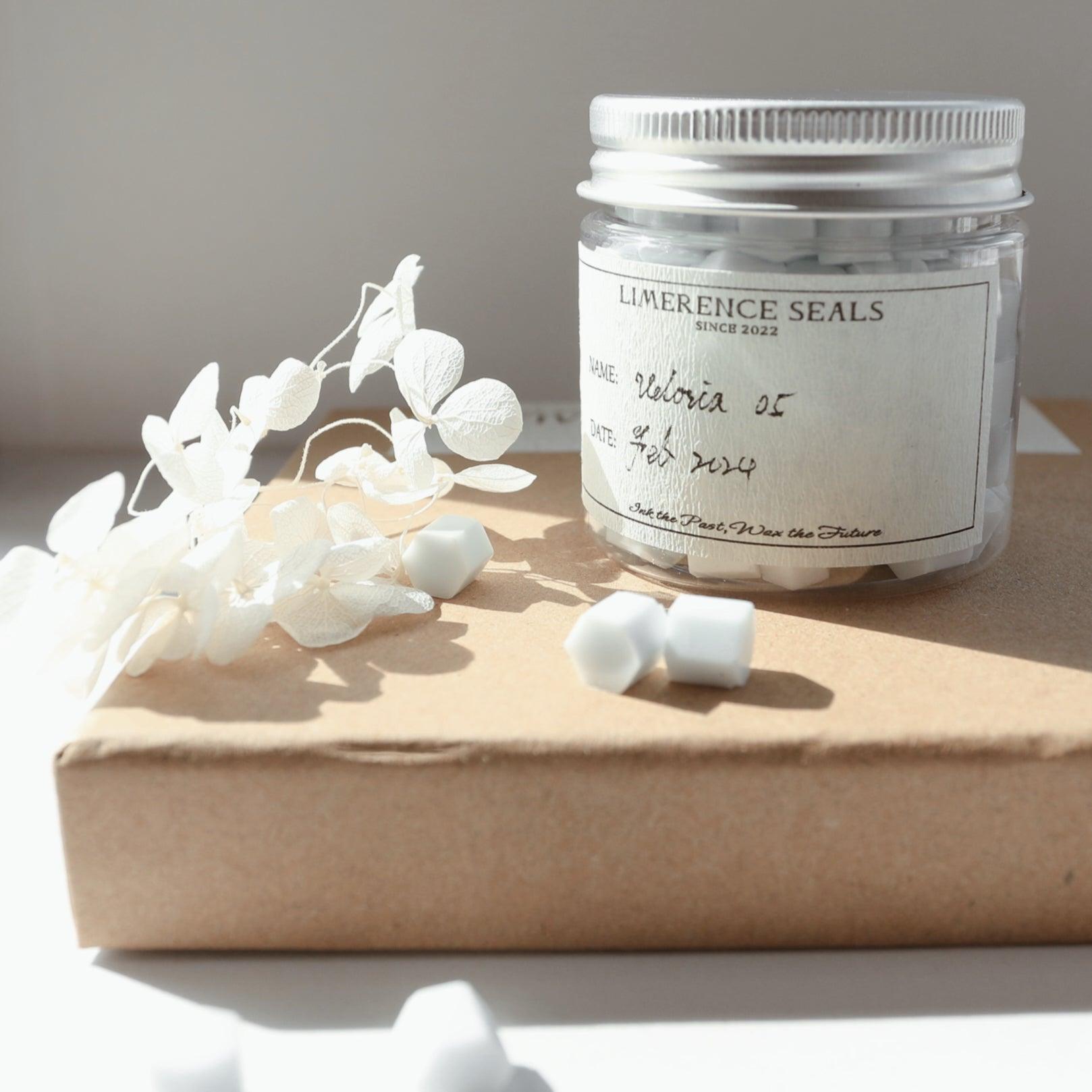 A jar of Veloria handmade sealing wax beads in light blue by Limerence Seals, placed on a kraft paper box. Delicate white flowers and scattered wax beads create a serene and elegant atmosphere, ideal for enhancing gift presentations and personalizing letters with a touch of tranquility and style.