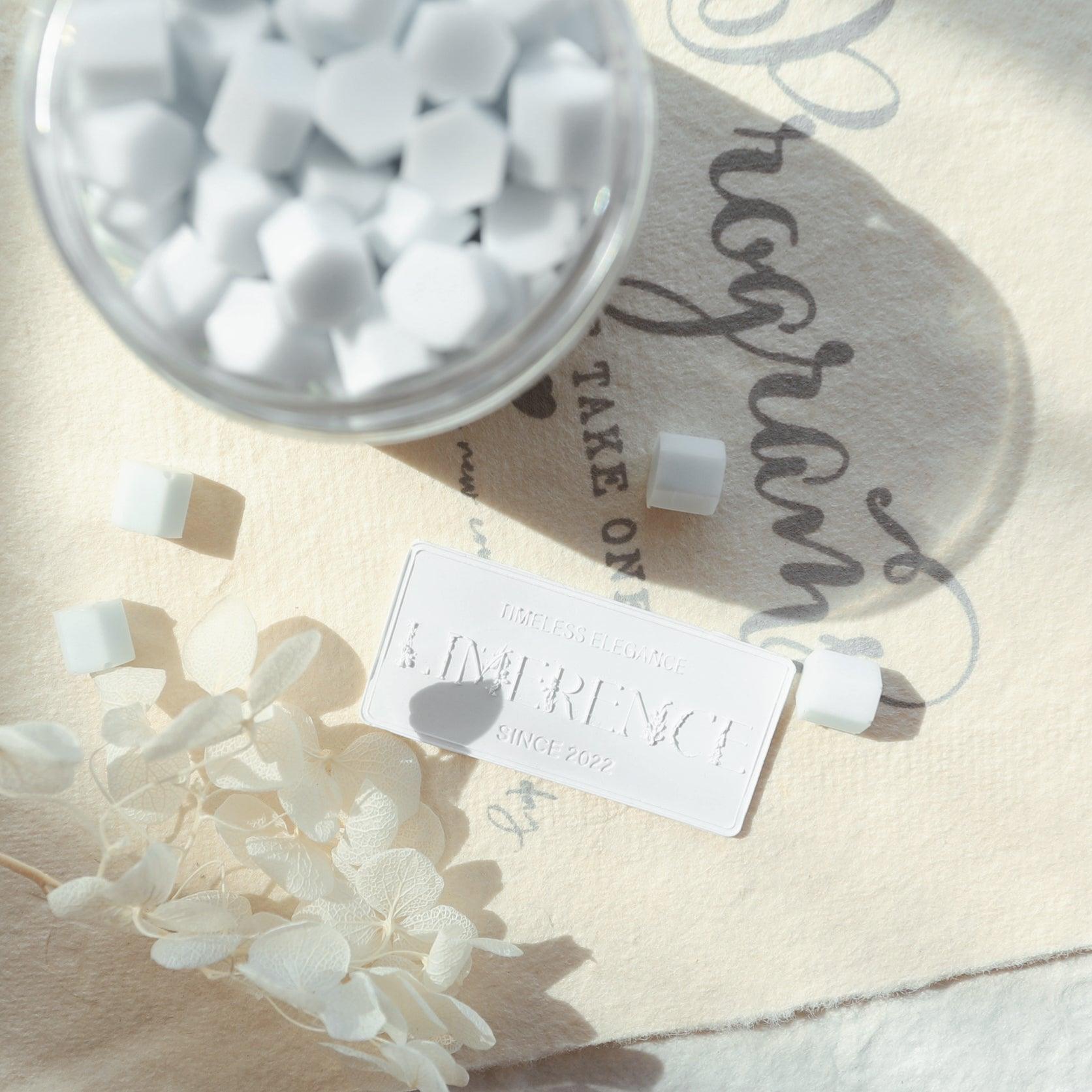 A jar of light blue Veloria handmade sealing wax beads by Limerence Seals, surrounded by a Limerence Seals label embossed with "TIMELESS ELEGANCE - LIMERENCE - SINCE 2022." The scene is completed with delicate white flowers on a fabric background with elegant script, highlighting the product's refined beauty and suitability for personalizing letters and special gifts.