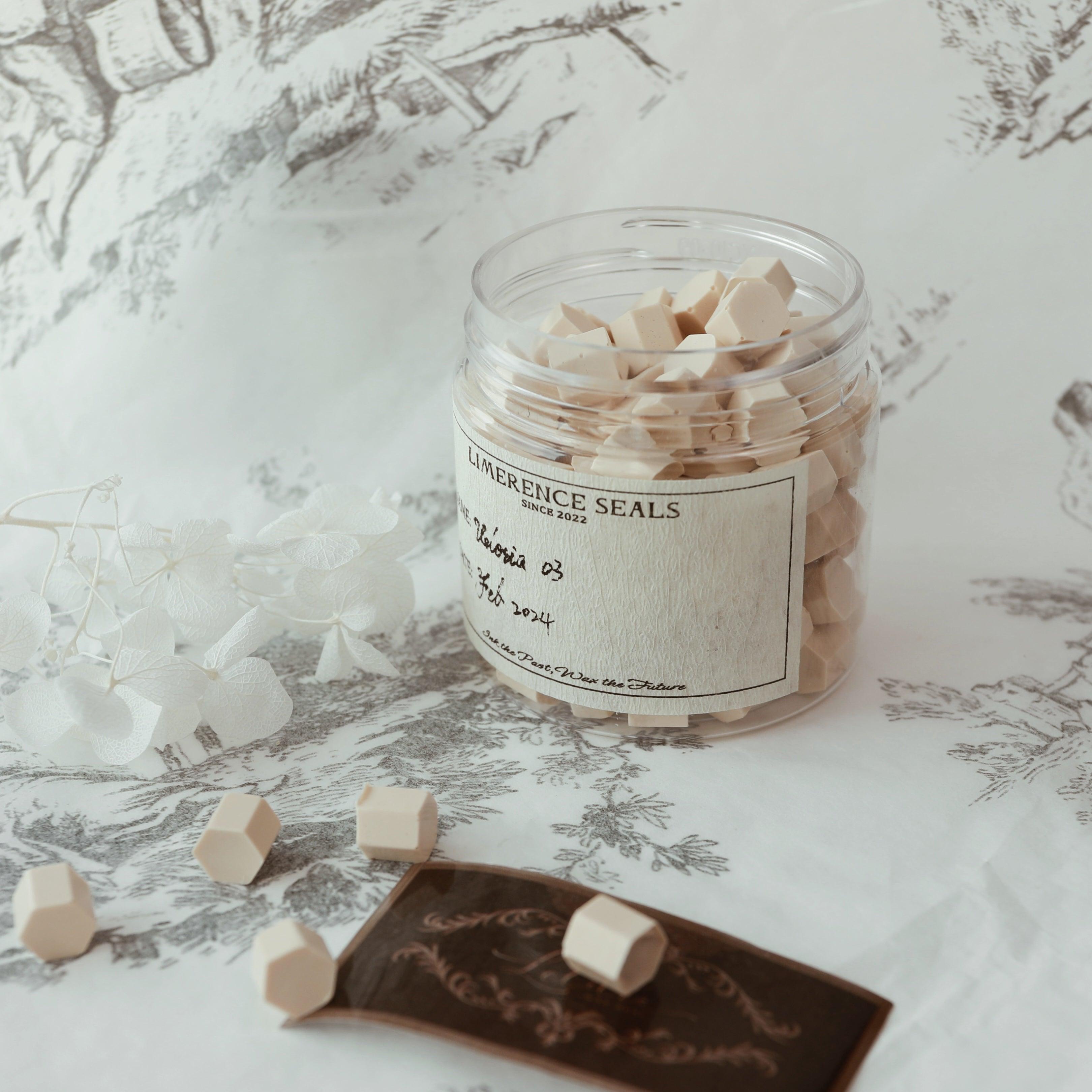 A jar of Veloria handmade sealing wax beads in light coffee color by Limerence Seals, set against an elegant monochrome floral background. Scattered beads and a decorative card emphasize the artistic and refined nature of the product, ideal for crafting exquisite seals on letters and invitations, reflecting sophistication and elegance.