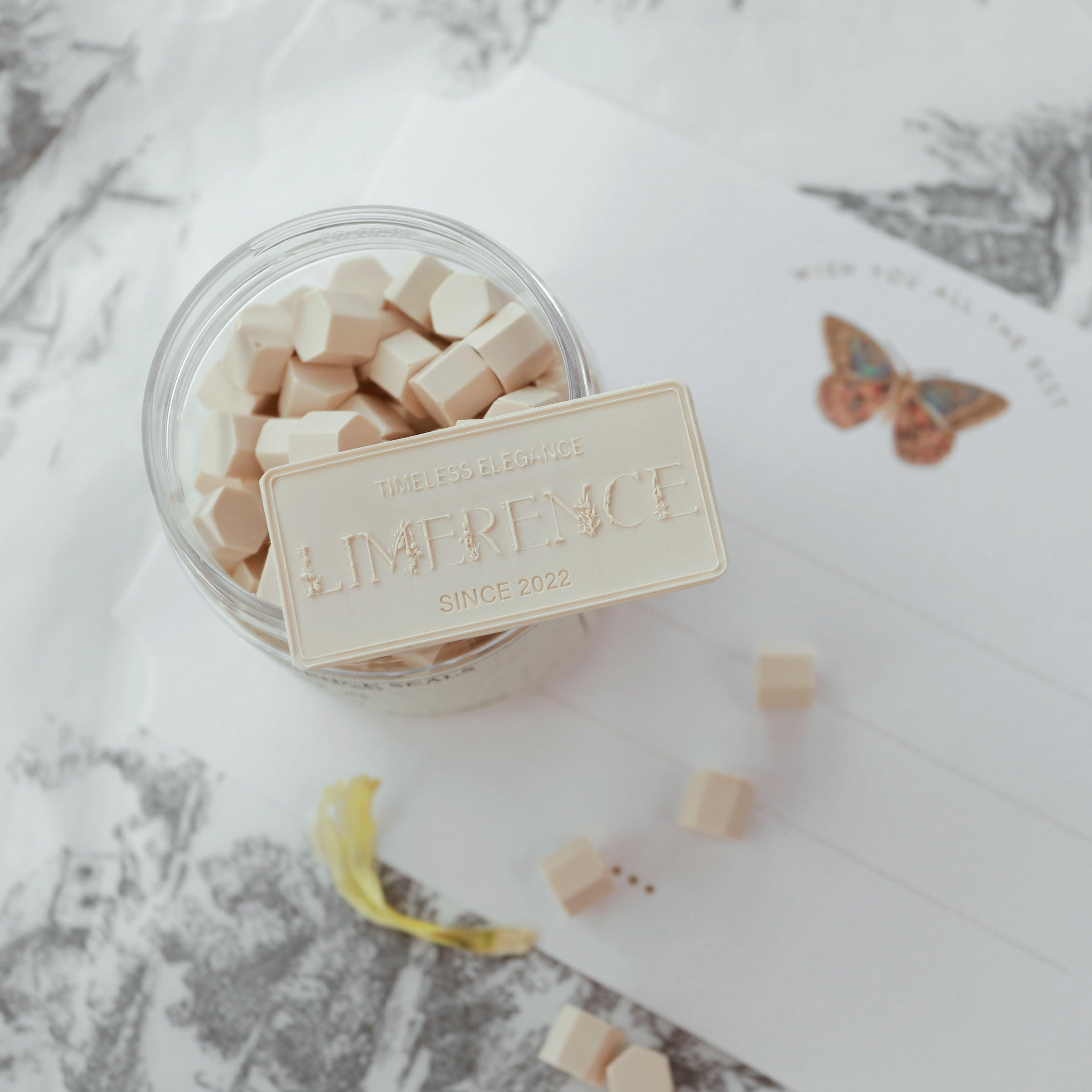 A jar of light coffee-colored Veloria handmade sealing wax beads by Limerence Seals, with a decorative label embossed with "TIMELESS ELEGANCE - LIMERENCE - SINCE 2022." The scene includes a card with a delicate butterfly illustration and scattered wax beads, highlighting the product's elegance and suitability for enhancing personalized letters and invitations.