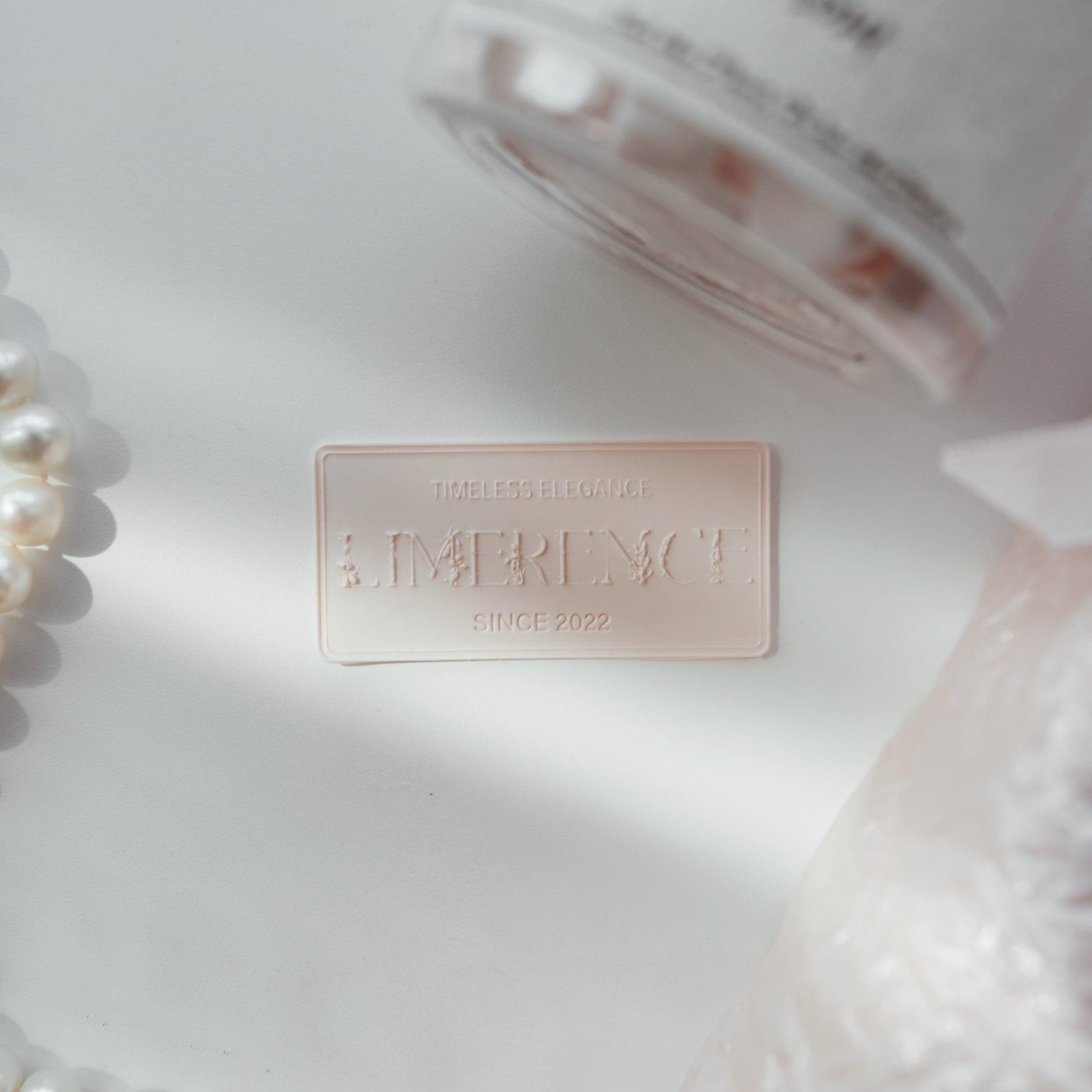 A light pink Limerence Seals label embossed with "TIMELESS ELEGANCE - LIMERENCE - SINCE 2022," elegantly placed on a white surface beside a string of pearls. The scene includes a jar of light pink Veloria handmade sealing wax beads, capturing a sense of delicate sophistication, perfect for upscale stationery and gift presentations.