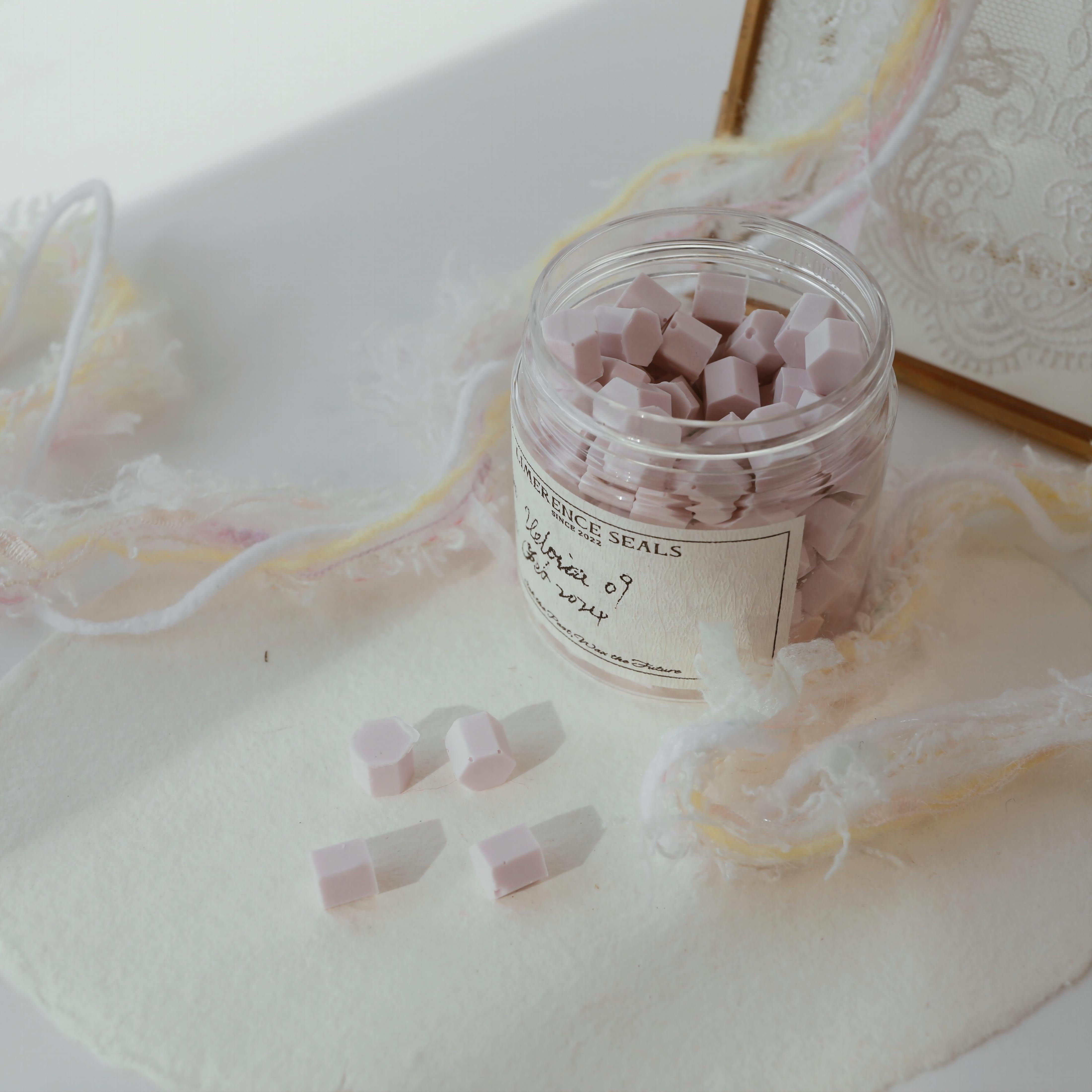 A jar of light purple Veloria handmade sealing wax beads by Limerence Seals, set on a soft fabric with pastel accents and surrounded by delicate lace. The gentle hue of the beads and the intricate fabric details create an elegant and whimsical aesthetic, perfect for adding a touch of charm to personalized letters and sophisticated gift packaging.