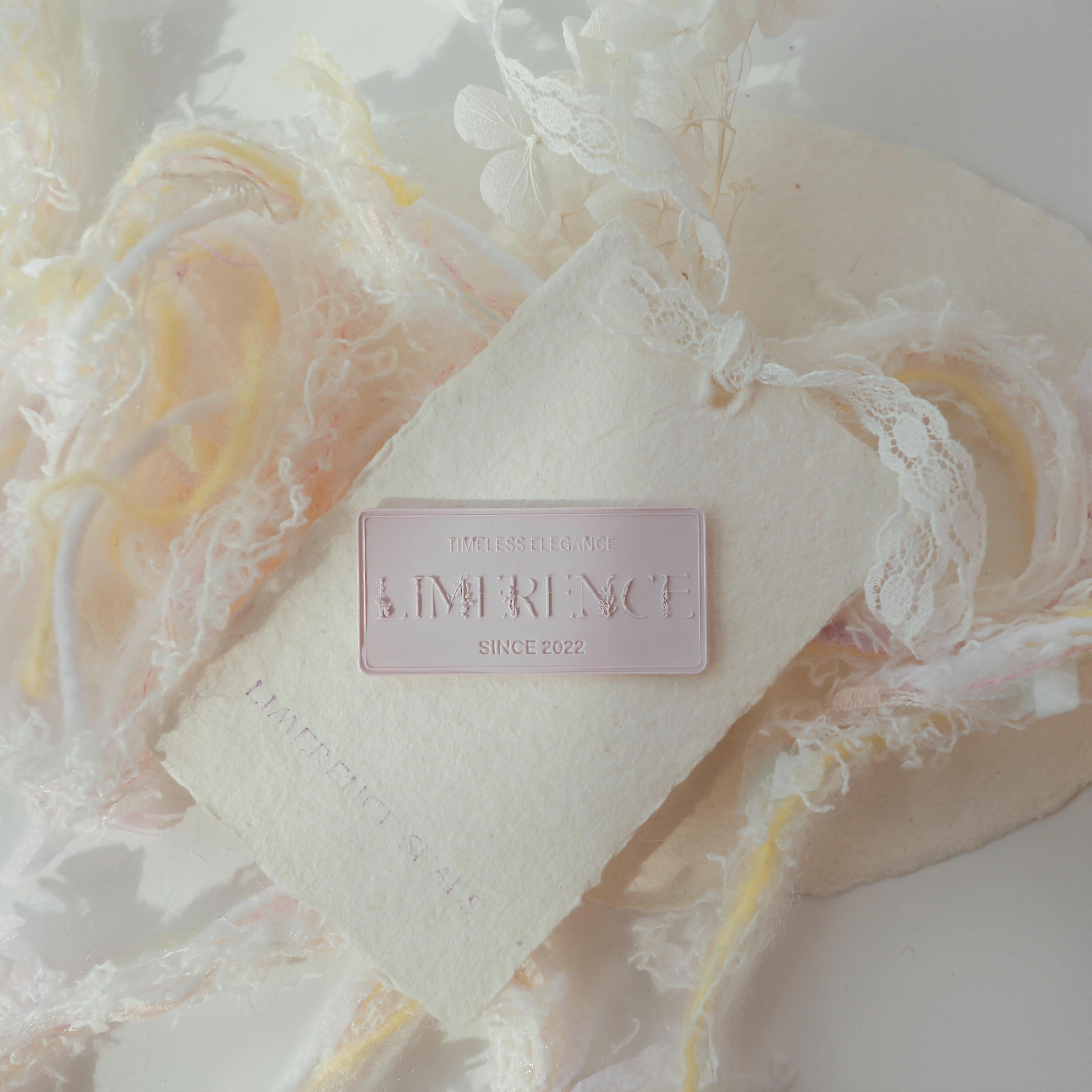 A light purple Limerence Seals label embossed with "TIMELESS ELEGANCE - LIMERENCE - SINCE 2022," placed on textured paper surrounded by soft pastel yarn and delicate lace. The combination of gentle colors and intricate textures creates a dreamy and sophisticated aesthetic, perfect for enhancing invitations and luxurious gift presentations.