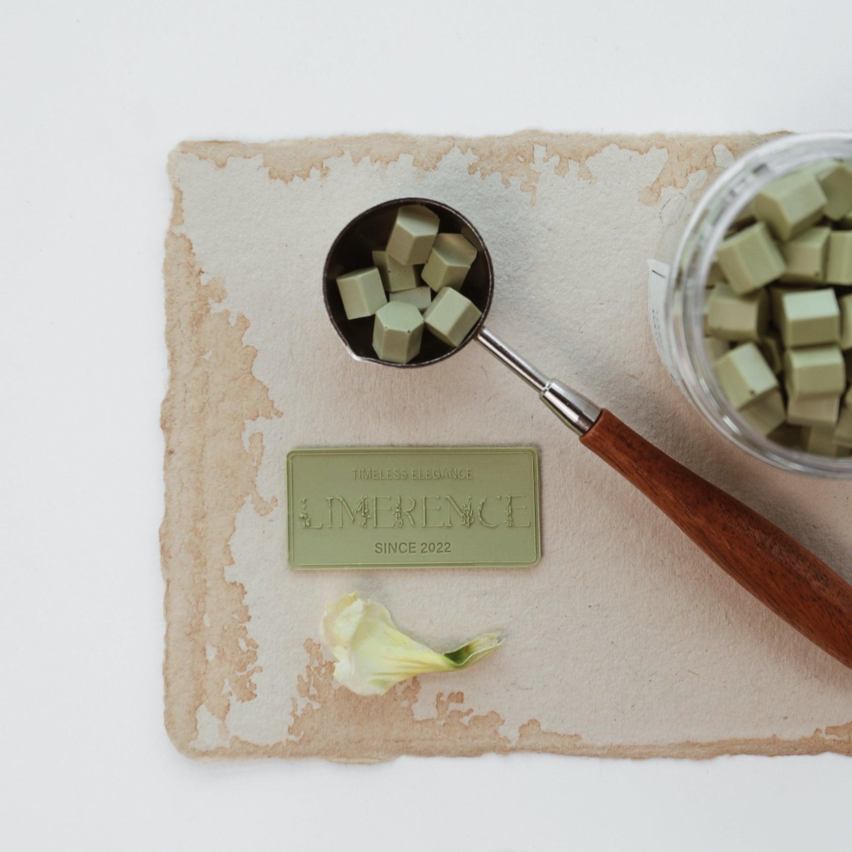 A matcha green Limerence Seals label embossed with "TIMELESS ELEGANCE - LIMERENCE - SINCE 2022" is set on textured parchment paper. Accompanied by a wooden-handled wax spoon filled with matcha green Veloria handmade sealing wax beads, the scene highlights a blend of natural elegance and traditional craftsmanship, ideal for enhancing letters and gift packaging.