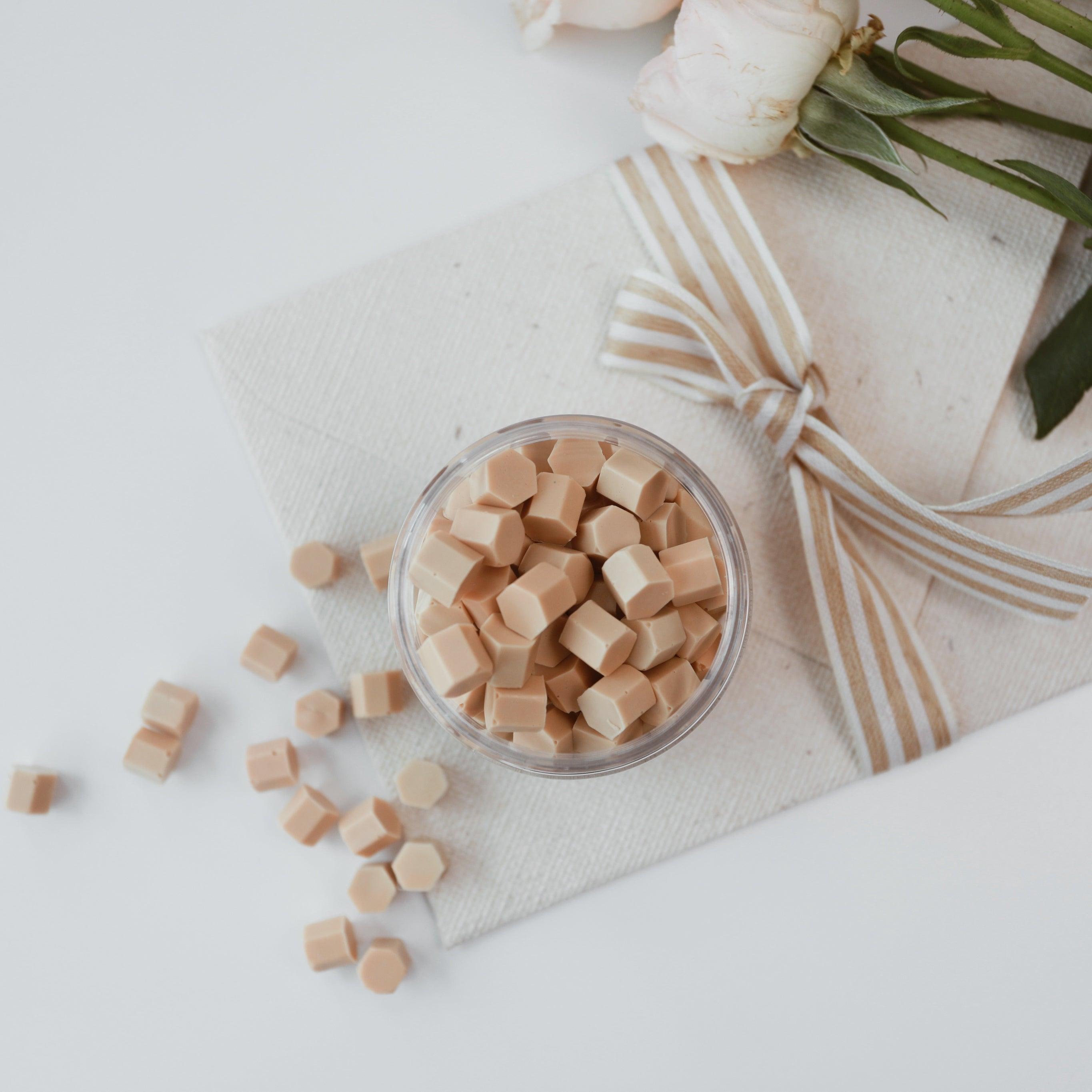 A jar of milk tea-colored Veloria handmade sealing wax beads by Limerence Seals, placed on a textured fabric envelope with a striped ribbon bow. Surrounding the setup are scattered wax beads and soft pink roses, showcasing the product's elegance and versatility for decorating invitations and enhancing special occasions with a touch of sophistication.