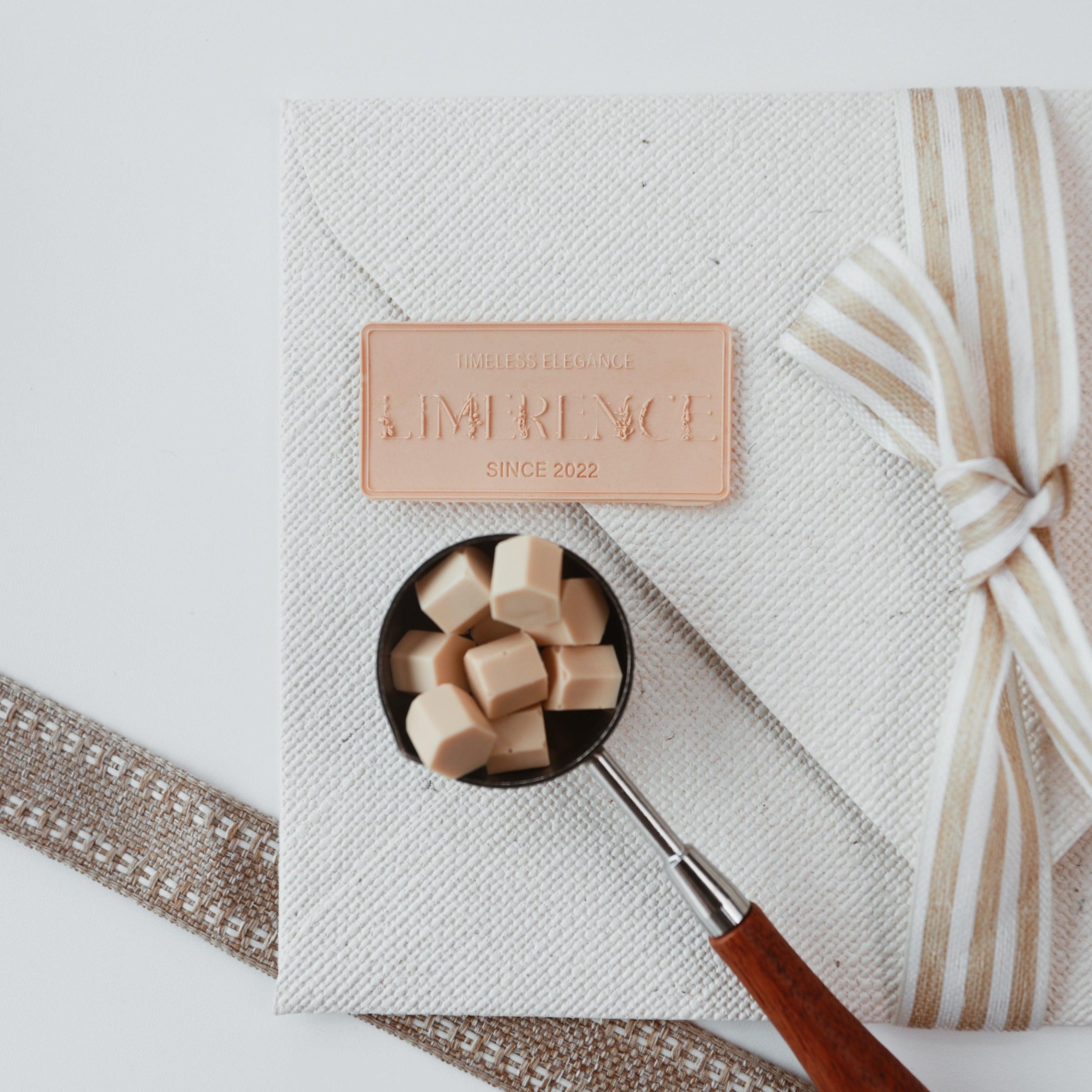 An elegant flat lay featuring a textured fabric envelope adorned with a milk tea-colored Limerence Seals label embossed with "TIMELESS ELEGANCE - LIMERENCE - SINCE 2022," accompanied by a striped ribbon bow. Beside it is a wax spoon with a wooden handle filled with milk tea-colored Veloria handmade sealing wax beads, showcasing the product's sophistication and ideal use for personalizing letters and gifts.