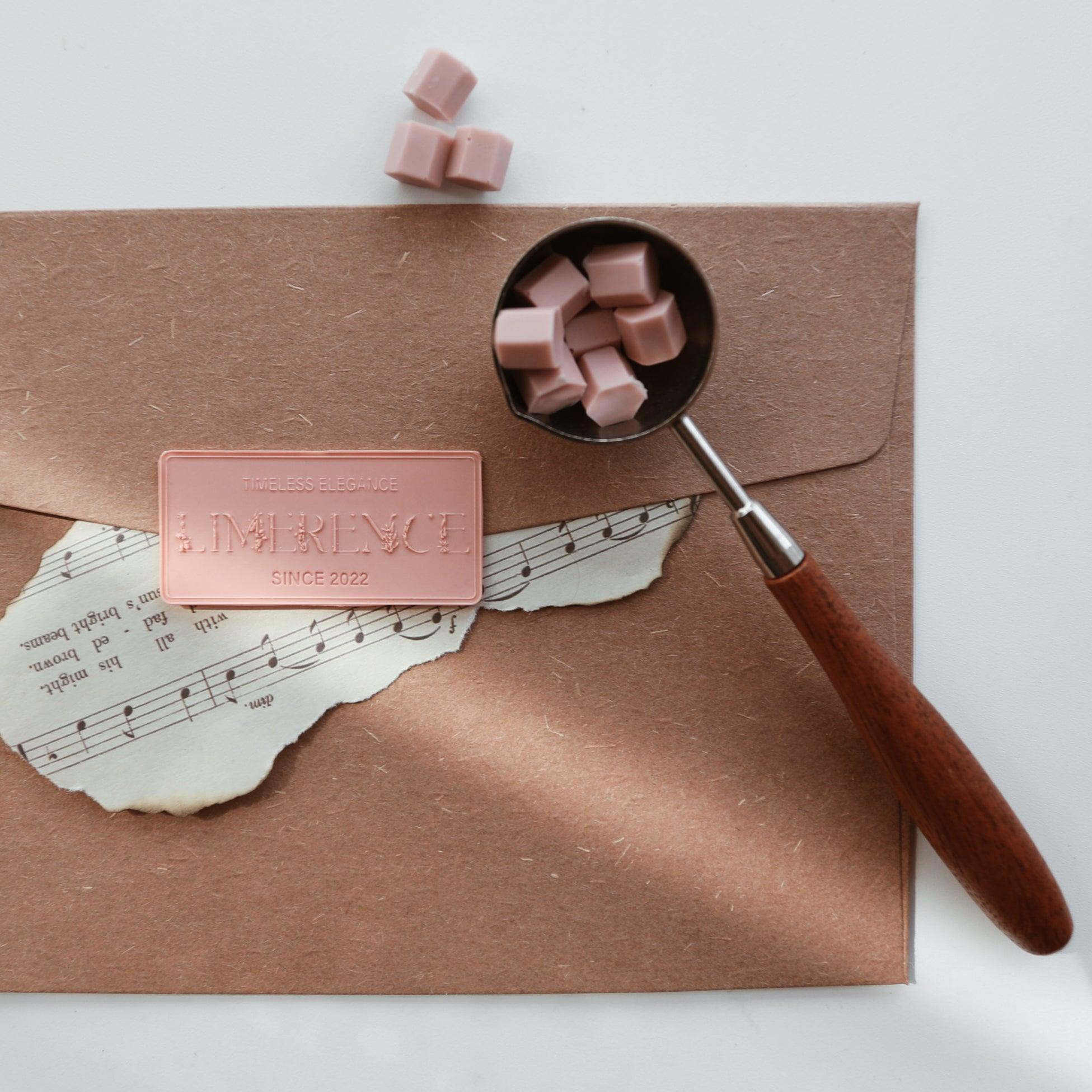 A scoop of peach pink Veloria handmade sealing wax beads by Limerence Seals resting on a brown paper envelope adorned with a torn sheet of vintage music paper and a Limerence label reading "TIMELESS ELEGANCE - LIMERENCE - SINCE 2022." The arrangement evokes a sense of nostalgia and elegance, perfect for adding a personal touch to letters and crafts.