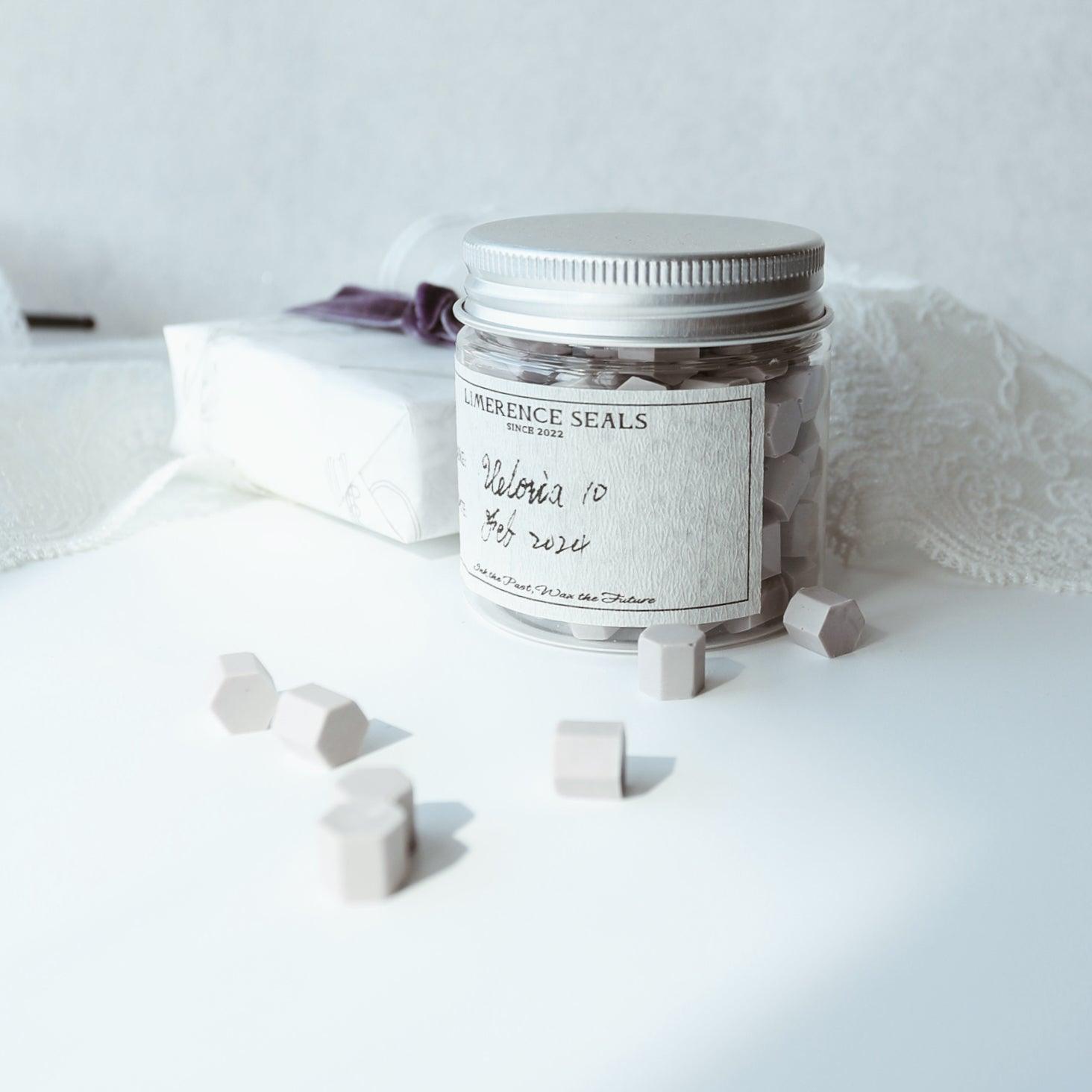 A jar of violet Veloria handmade sealing wax beads by Limerence Seals, set on a pristine white surface with a background of delicate lace and a wrapped gift. The soft lighting highlights the subtle purple tones of the beads, perfect for adding a touch of elegance to personal letters and refined gift presentations.